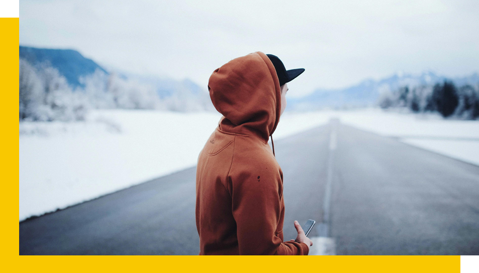 Person in a hooded jacket standing on an empty road with snowy landscape in the background.